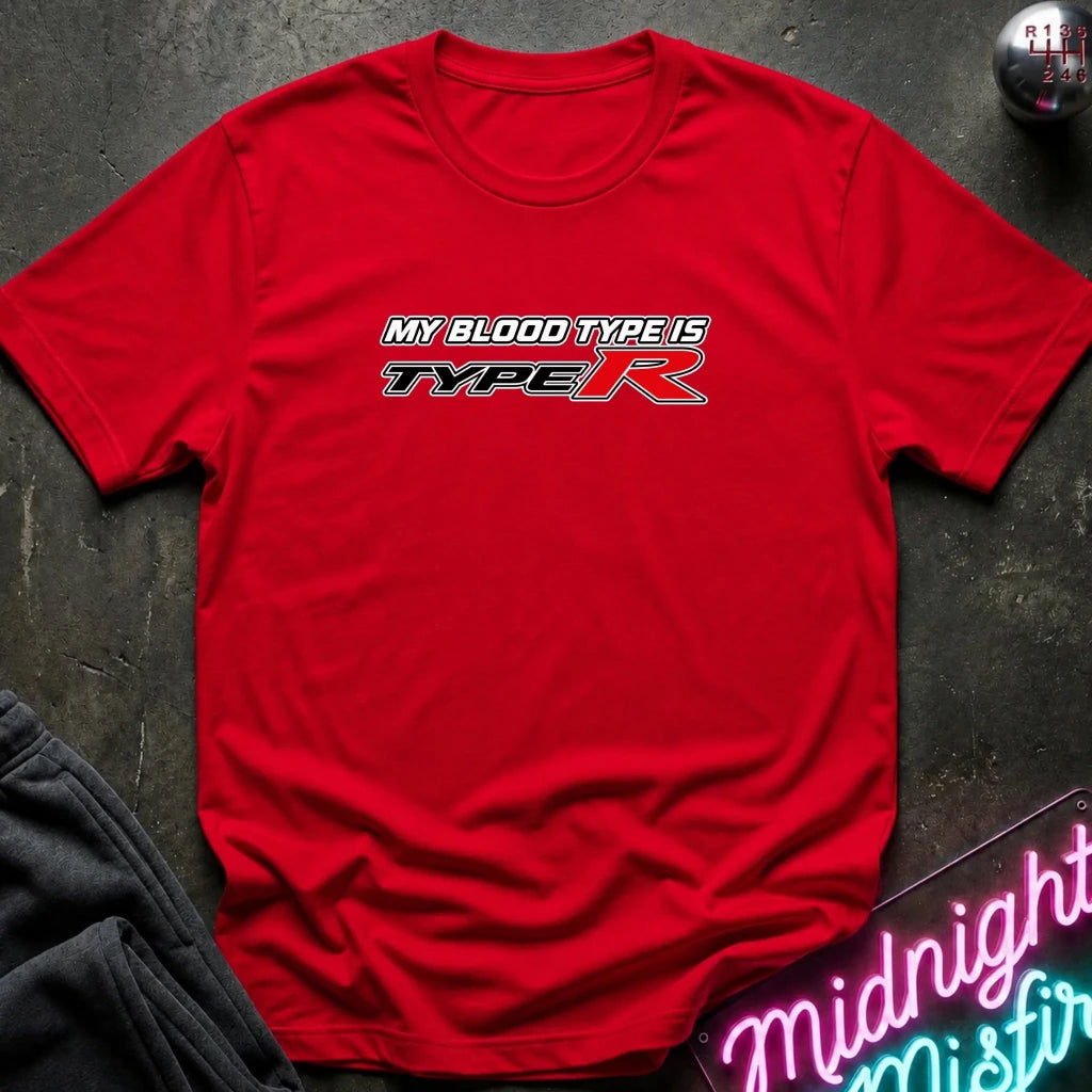 A red short-sleeve crewneck t-shirt with a 'MY BLOOD TYPE IS TYPE R' graphic printed on the chest, laid flat on a dark textured surface with a gear and neon sign props.