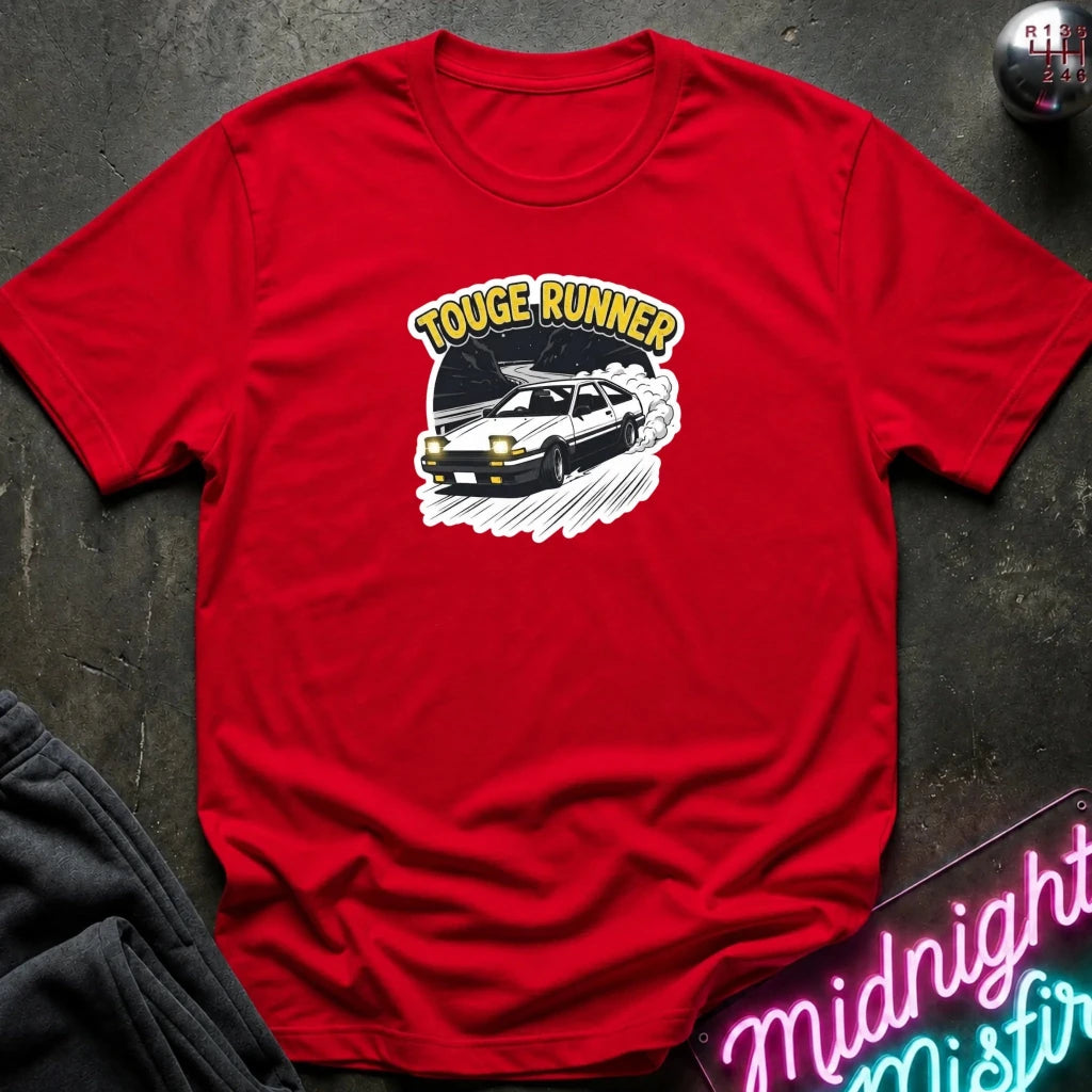 red short-sleeve t-shirt laid flat on a dark textured surface, featuring a graphic of a black sports car with the words 'TOUGE RUNNER' in yellow above it. A neon sign and folded dark fabric are partially visible around the edges.