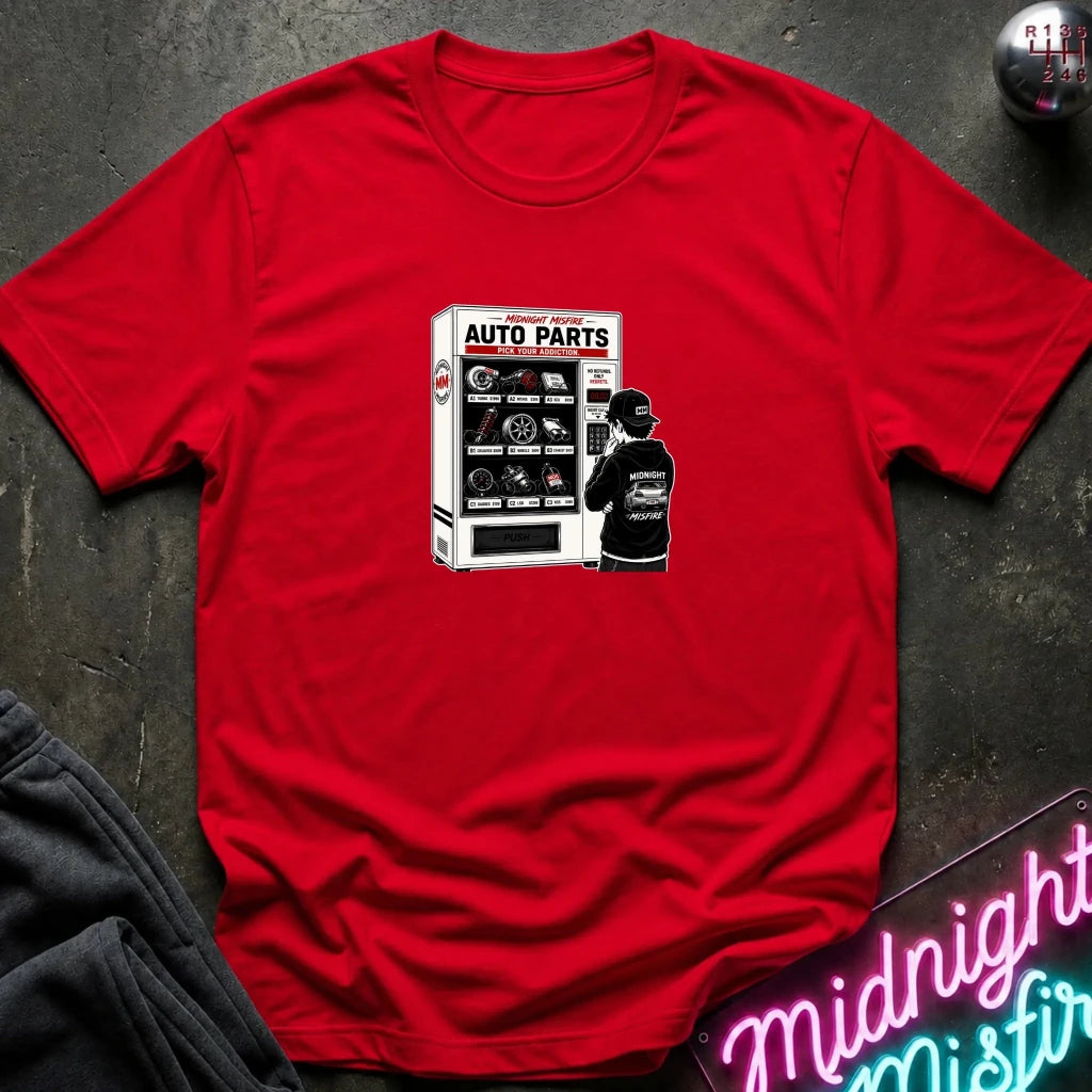 A red short-sleeve crewneck t-shirt laid flat on a dark textured surface, featuring a graphic print of a person standing in front of an 'Auto Parts' vending machine. 