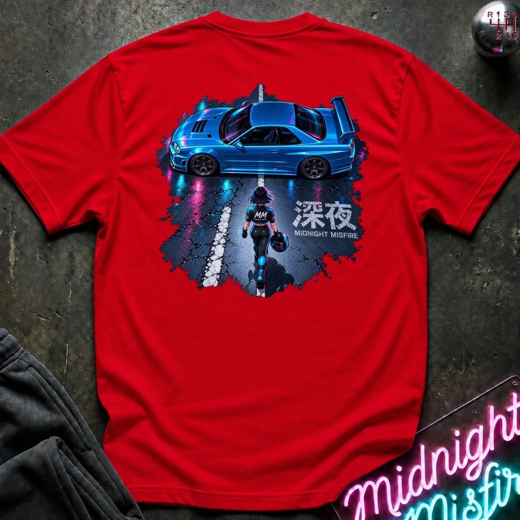 A red short-sleeve T-shirt laid flat on a dark textured surface, featuring a large back graphic of a blue sports car on a wet road with a motorcycle and Japanese text. 