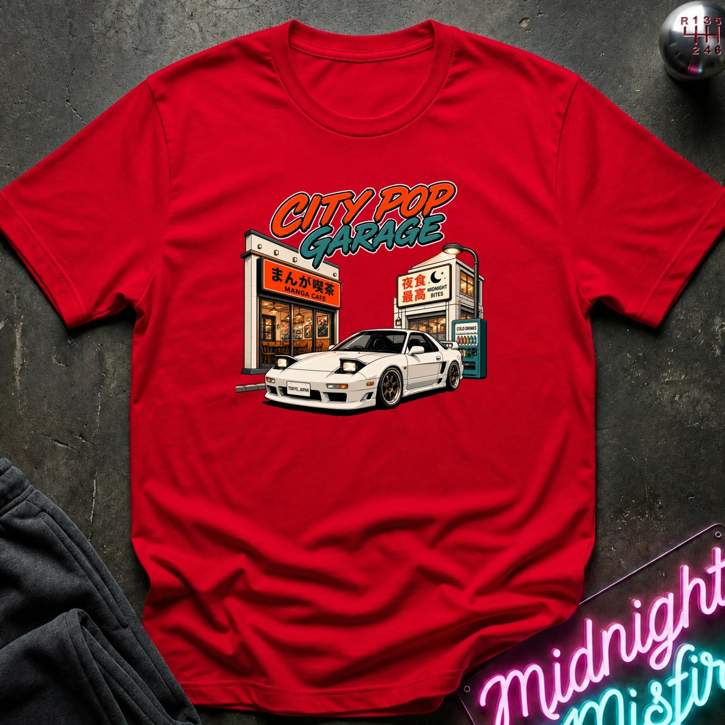 A red short-sleeve graphic t-shirt laid flat on a dark textured surface. The graphic features a white sports car parked in front of a cafe with the text 'CITY POP GARAGE' in orange and teal.