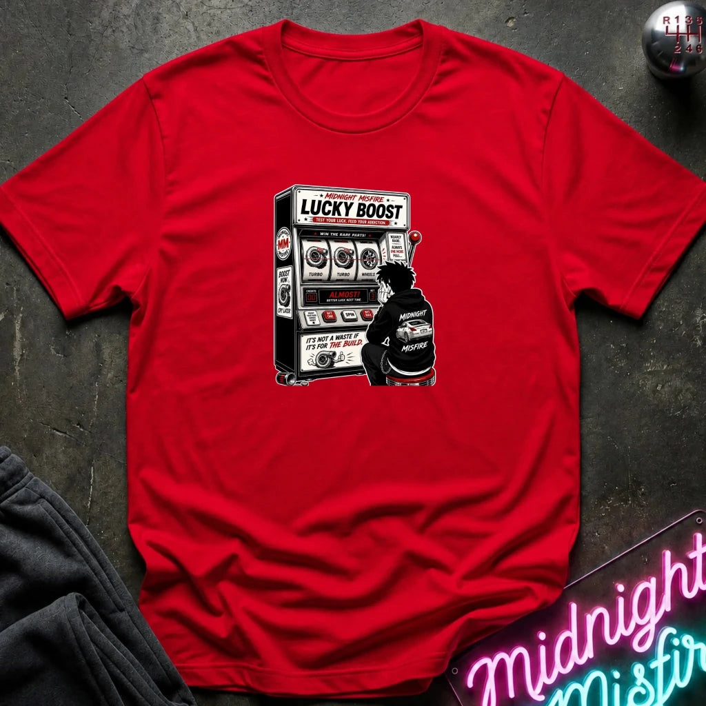 A red short-sleeve crewneck t-shirt laid flat on a dark textured surface, featuring a black and white graphic of a person sitting in front of a 'Lucky Boost' slot machine. Styled flat-lay props include a dark sweatshirt in the bottom-left corner and a pink neon sign in the bottom-right corner.