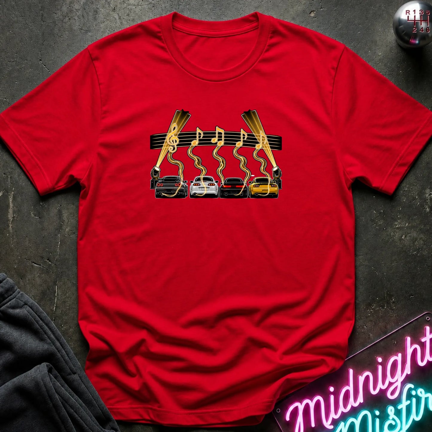 A red short-sleeve t-shirt laid flat on a dark textured surface, featuring a graphic print of four cars (red, white, gray, yellow) with gold wavy lines and musical notes above them, flanked by two  lights.