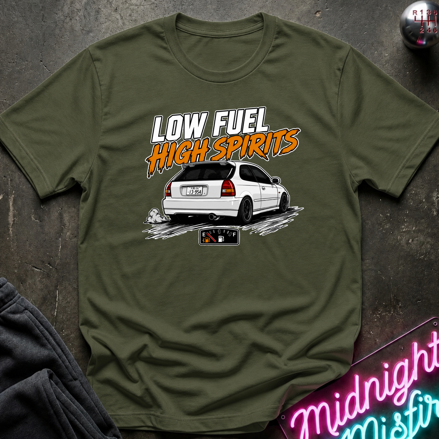 An olive green graphic t-shirt laid flat on a dark textured surface. The shirt features a white car graphic and the text 'LOW FUEL HIGH SPIRITS' in white and orange.