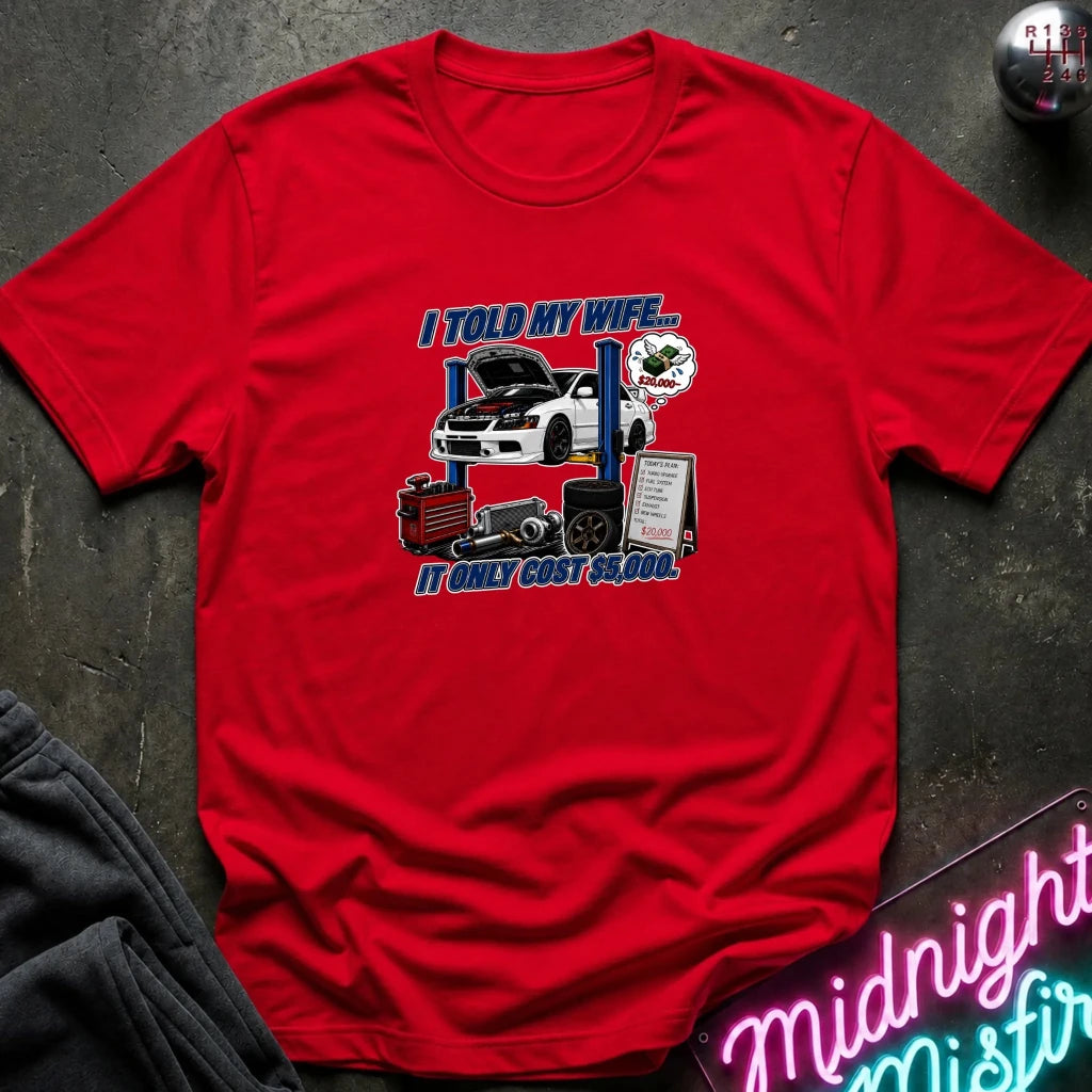 A red short-sleeve graphic t-shirt laid flat on a dark textured surface. The graphic features a white sports car on a blue lift with the text 'I TOLD MY WIFE.