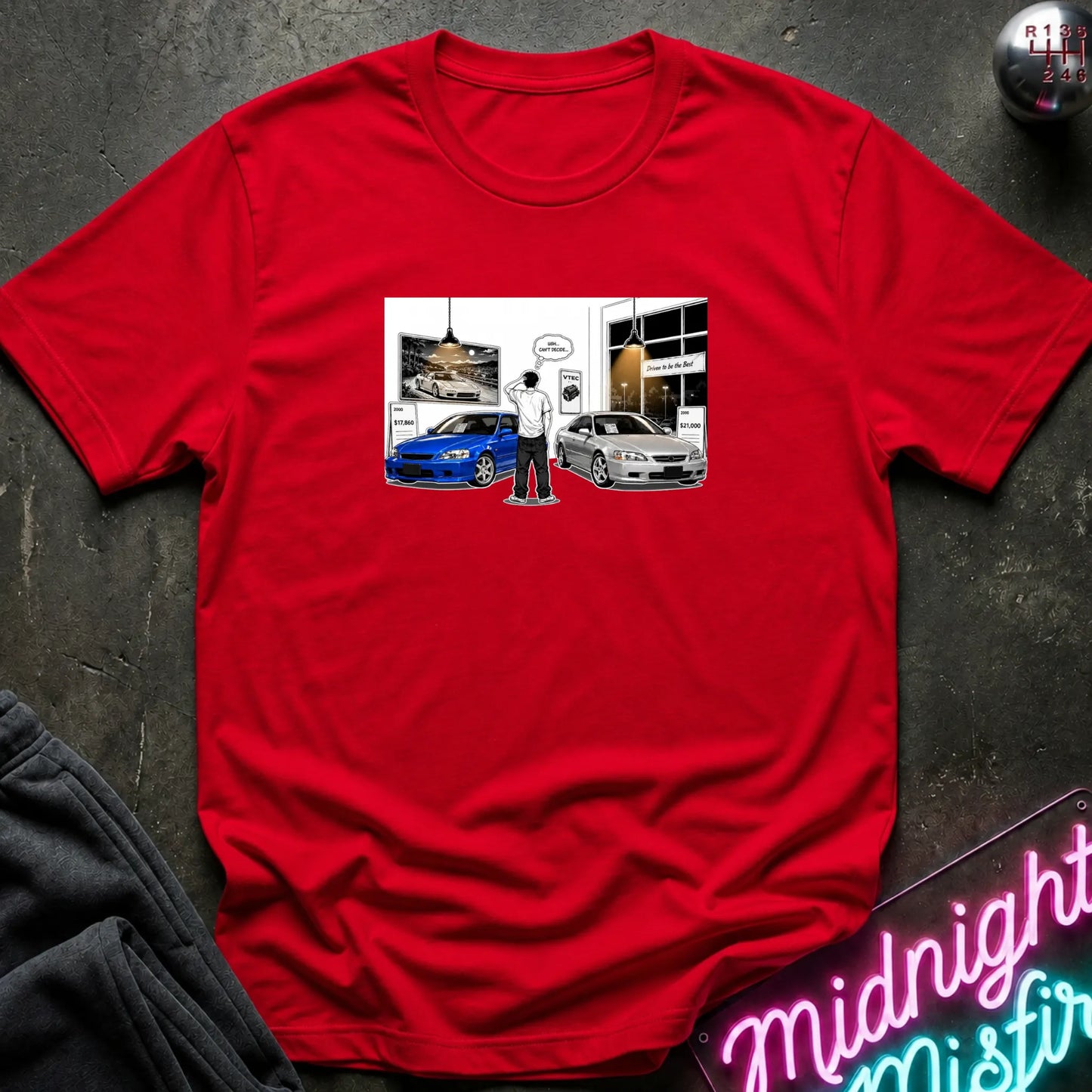 A red short-sleeve crewneck t-shirt laid flat on a dark textured surface. The center of the shirt features a rectangular graphic print depicting a cartoon character standing between a blue sports car and a silver sedan in front of a building.