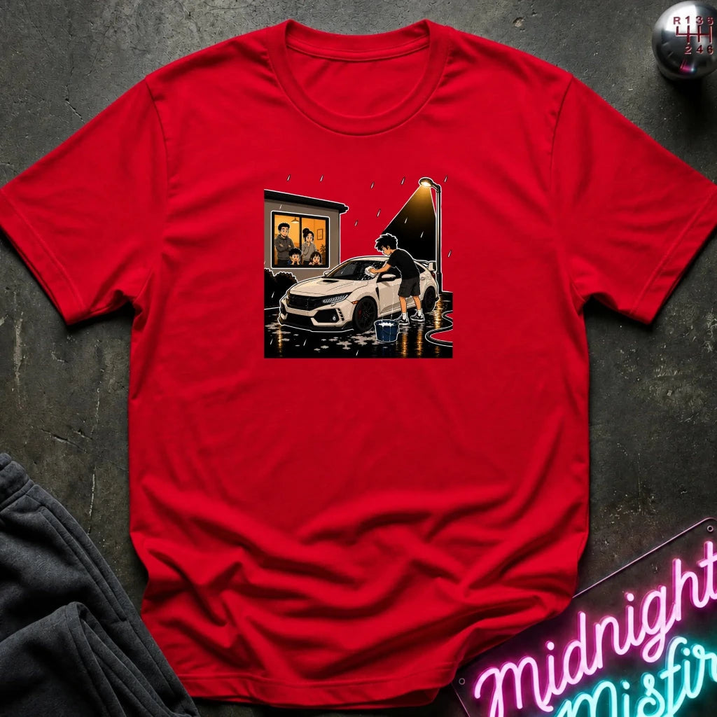 A red short-sleeve crewneck t-shirt laid flat on a dark textured surface, featuring a central graphic print of a white sports car in a rainy street scene. A gray sweatshirt corner and a neon sign are partially visible as styling props.