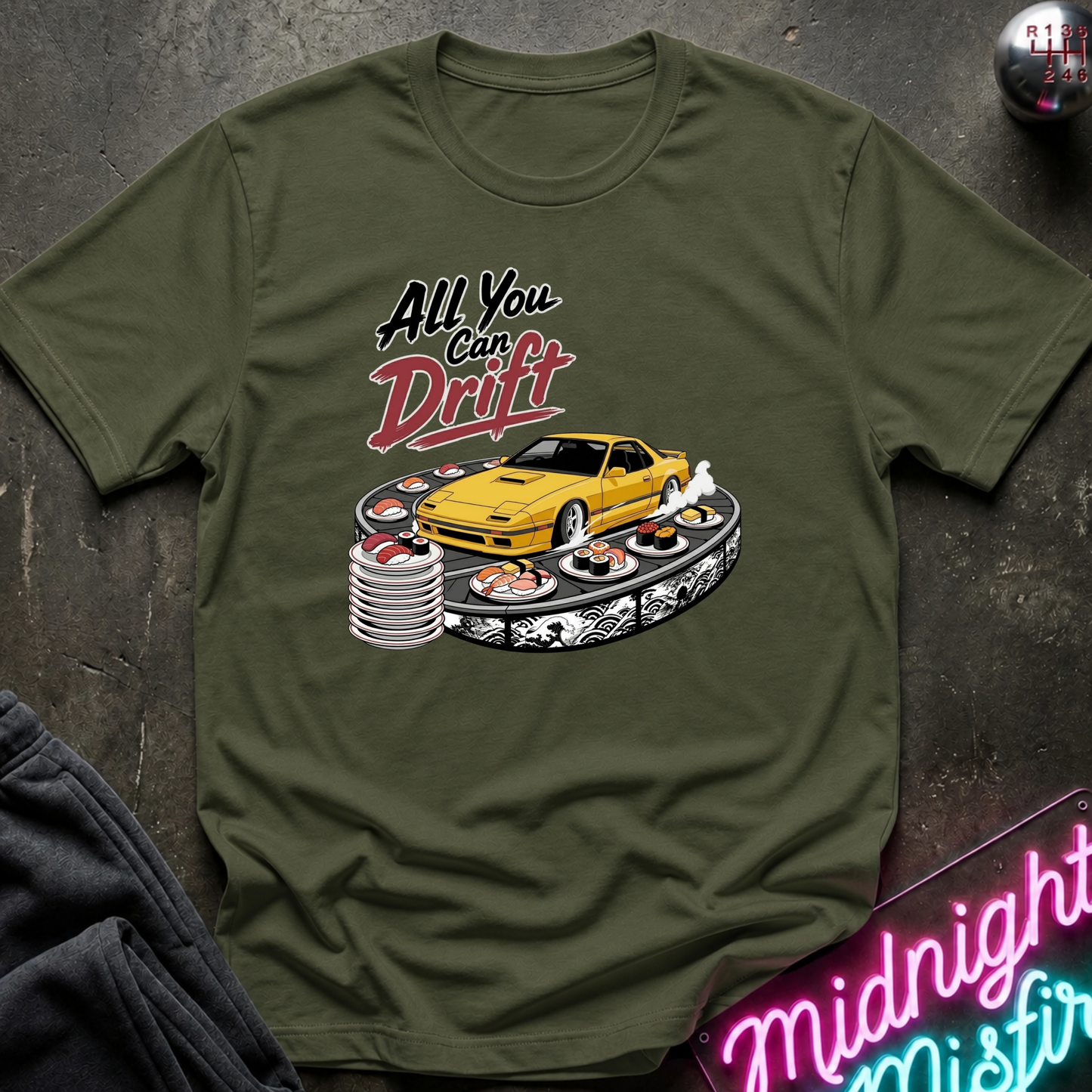 An olive green graphic t-shirt laid flat on a dark textured surface. The graphic features a yellow sports car drifting on a track with the text 'All You Can Drift' in stylized black and red lettering.