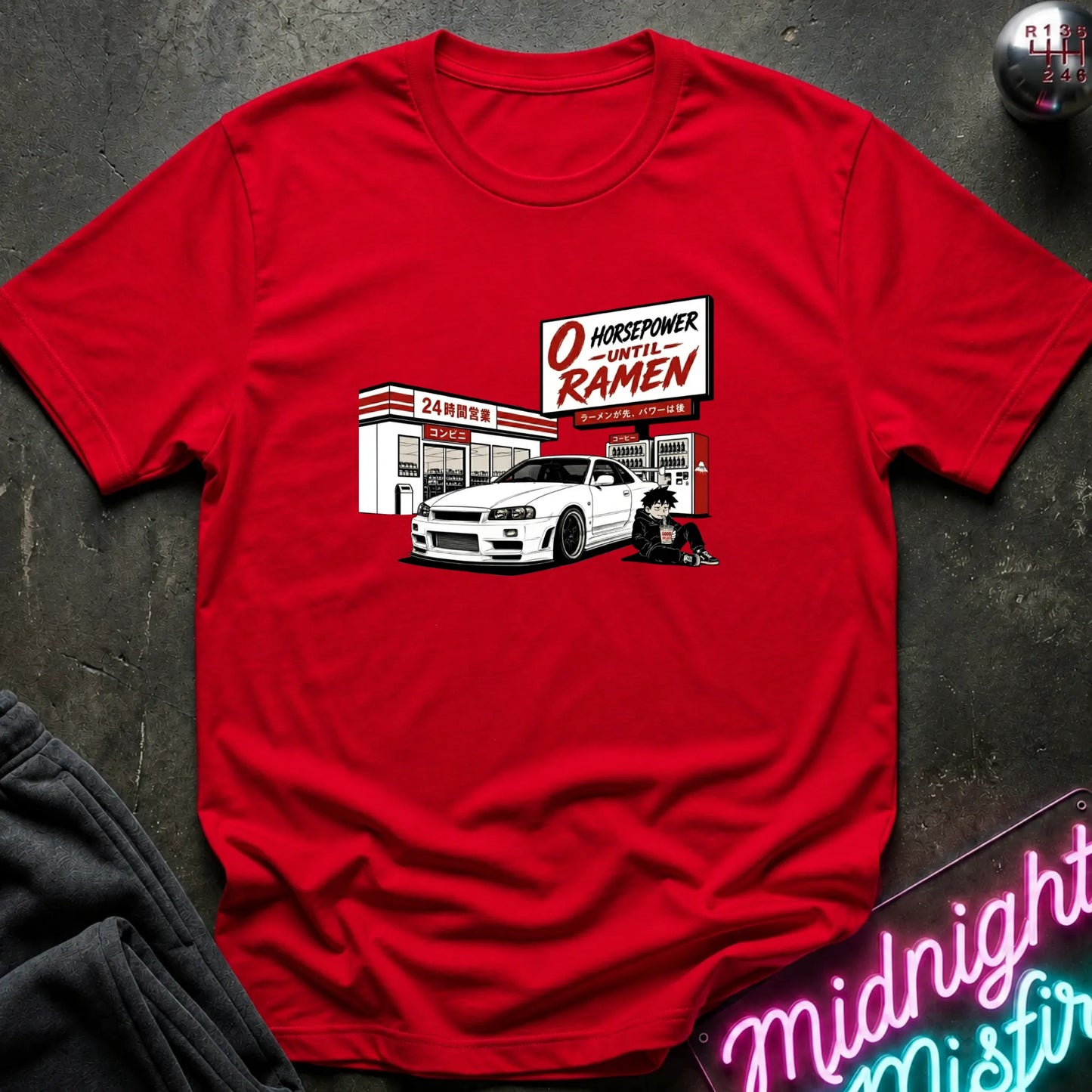 A red short-sleeve crewneck t-shirt featuring a graphic print of a white sports car parked in front of a store with a sign reading 'O HONORPOWER -UNTIL RAMEN'. The shirt is displayed flat on a dark textured surface with a neon sign and dark fabric partially visible in the corners.