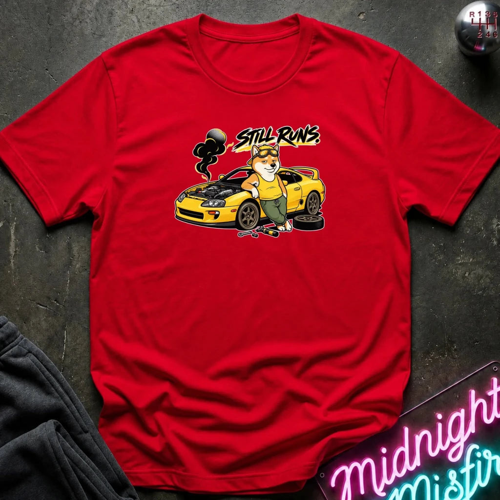 red short-sleeve graphic t-shirt laid flat on a dark textured surface. The graphic features a cartoon dog in a mechanic hat standing next to a yellow sports car with the text 'Still runs' above it.