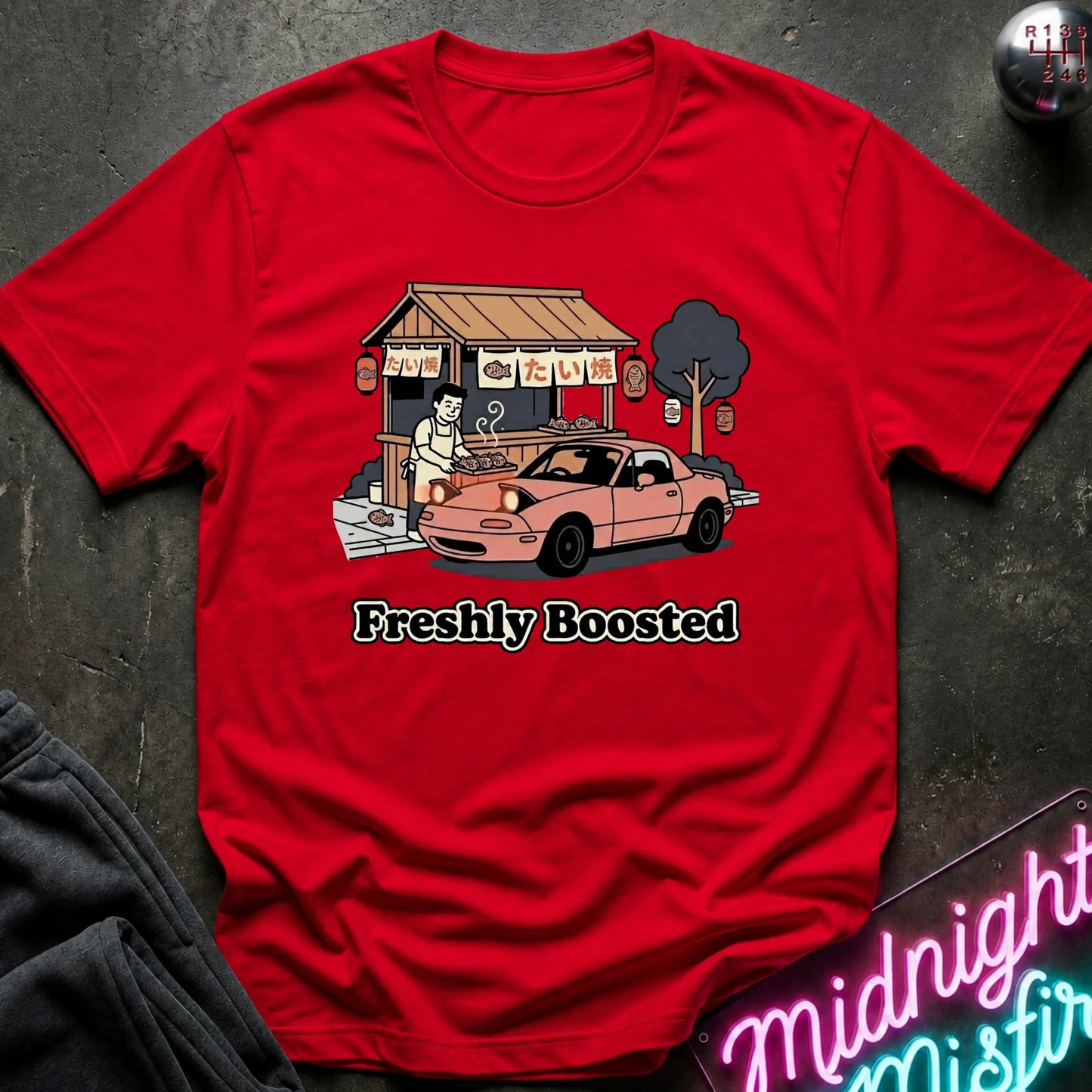 A red short-sleeve graphic T-shirt laid flat on a dark textured surface. The shirt features a cartoon illustration of a street food stall with a pink car and the text 'Freshly Boosted' below it.