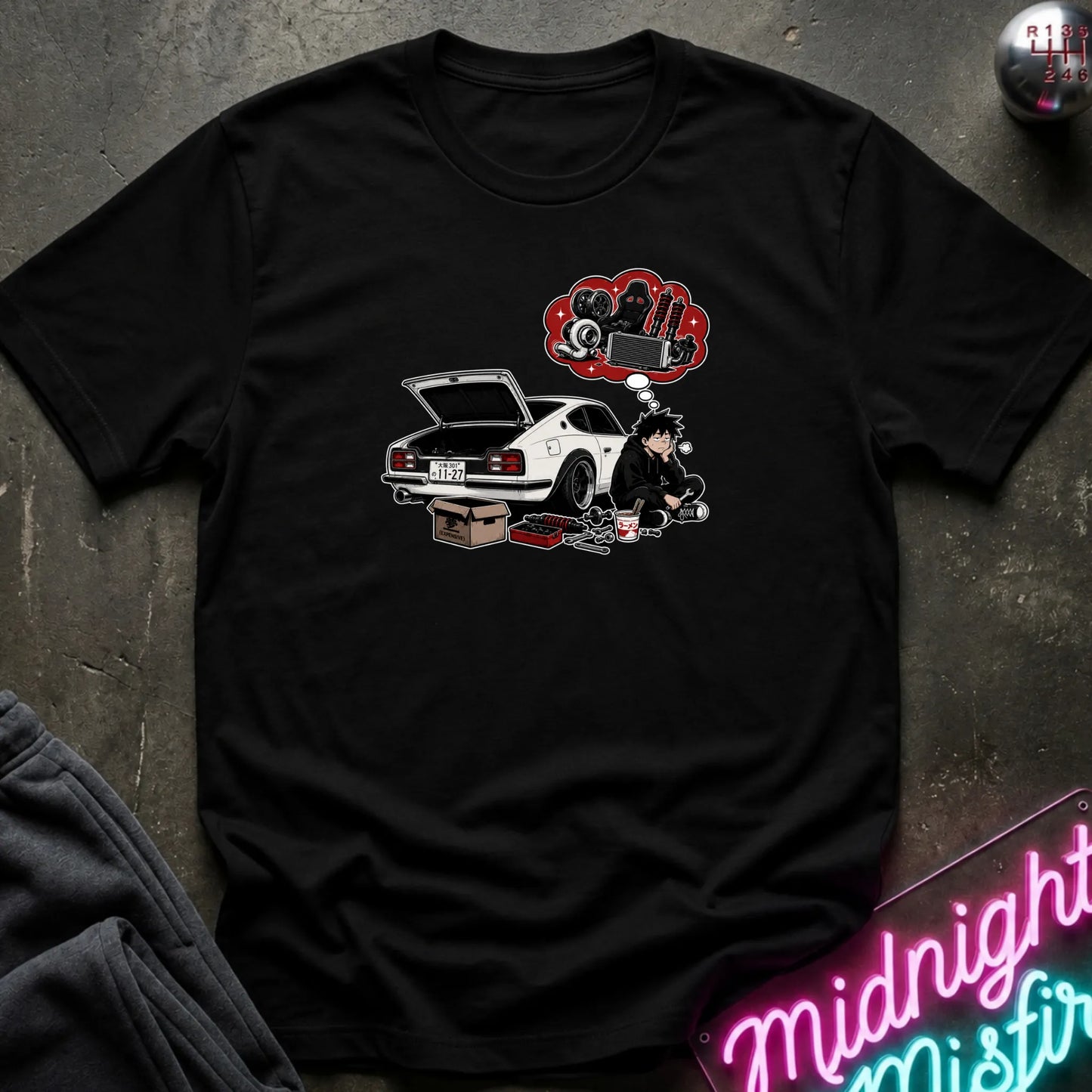 A black short-sleeve graphic t-shirt laid flat on a dark textured surface. The graphic features a white sports car with its trunk open, a cartoon character sitting beside it, and a red thought bubble filled with car parts.