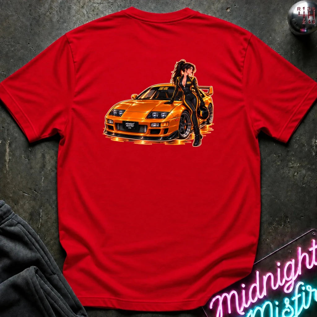 A red short-sleeve t-shirt laid flat on a dark textured surface, featuring a large graphic print of an orange sports car with a woman in a black outfit leaning on it. A gear shift knob is visible in the top right corner, and a neon sign is partially visible in the bottom right.