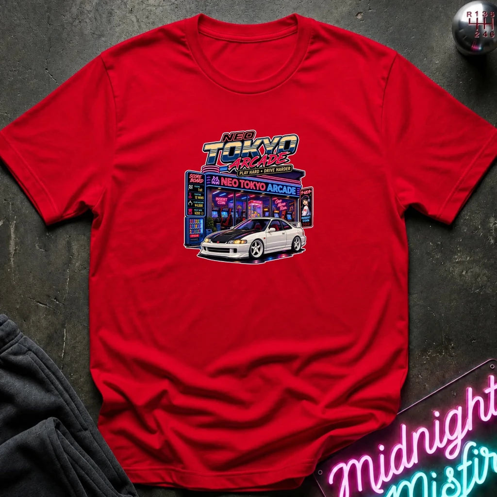 A red short-sleeve crewneck t-shirt featuring a 'Neo Tokyo Arcade' graphic with a white sports car, laid flat on a dark textured surface with a neon sign and sweatpants as props.