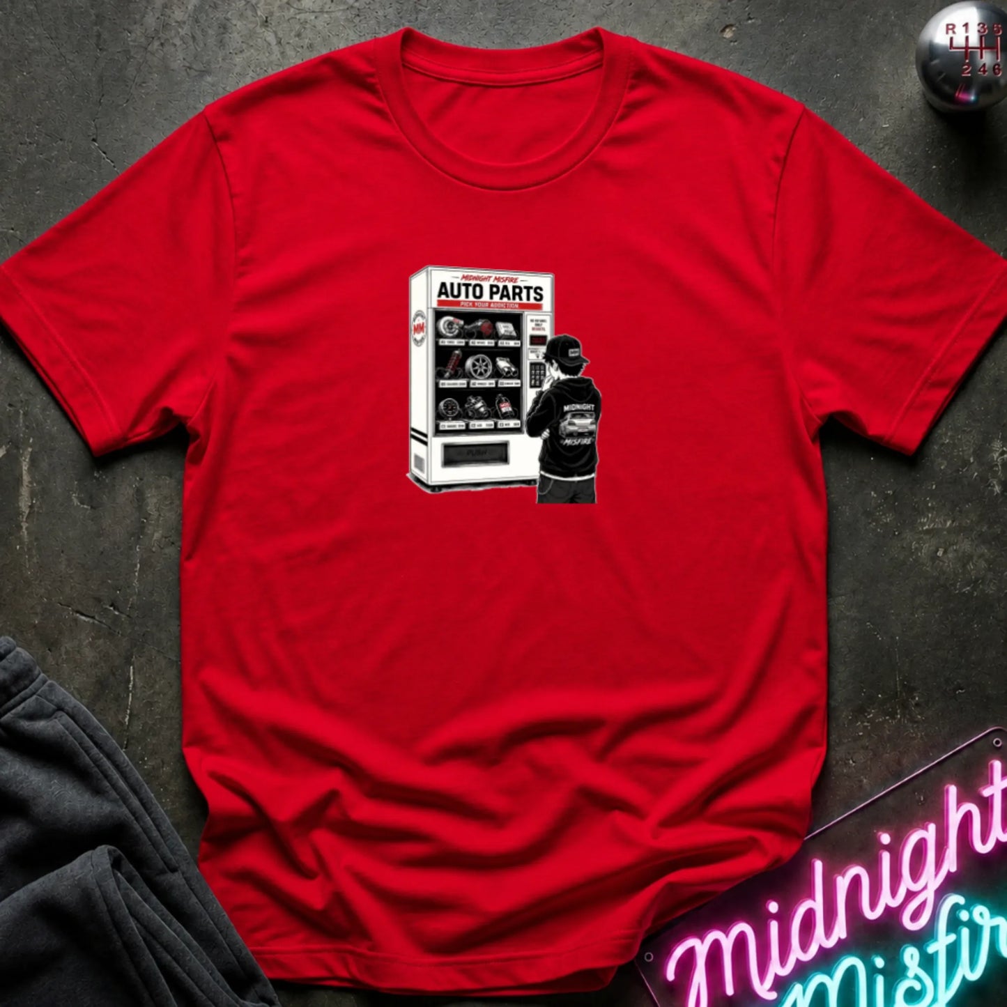 A red short-sleeve t-shirt laid flat on a dark textured surface, featuring a graphic print of a vending machine and owner deciding.
