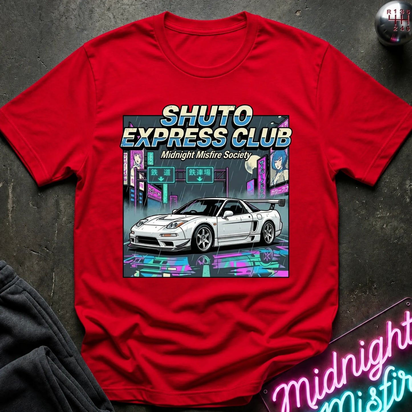 A red short-sleeve graphic T-shirt laid flat on a dark textured surface. The shirt features a large front print with the words “SHUTO EXPRESS CLUB” and “Midnight Misfire Society” above an illustration of a white sports car in a neon cyberpunk city scene.