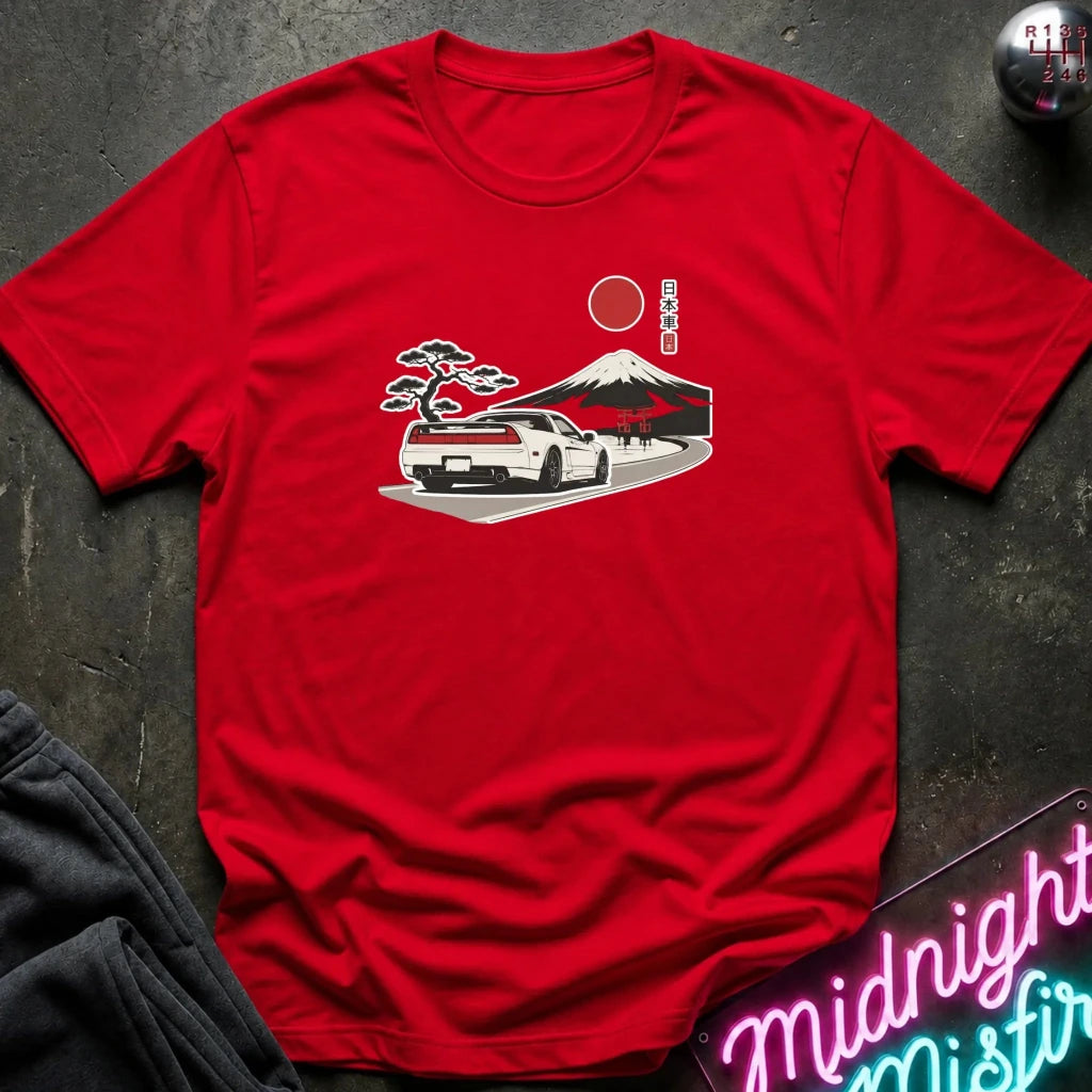 A red short-sleeve t-shirt laid flat on a dark textured surface, featuring a graphic print of a white sports car on a road with a red sun, Mount Fuji, and a pine tree in a Japanese-style illustration.