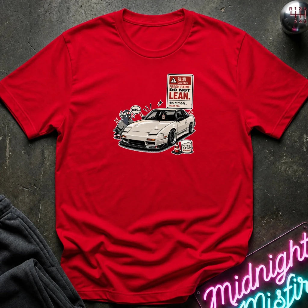 A red short-sleeve t-shirt laid flat on a dark textured surface, featuring a graphic print of a white sports car next to a 'DO NOT LEAN' warning sign.