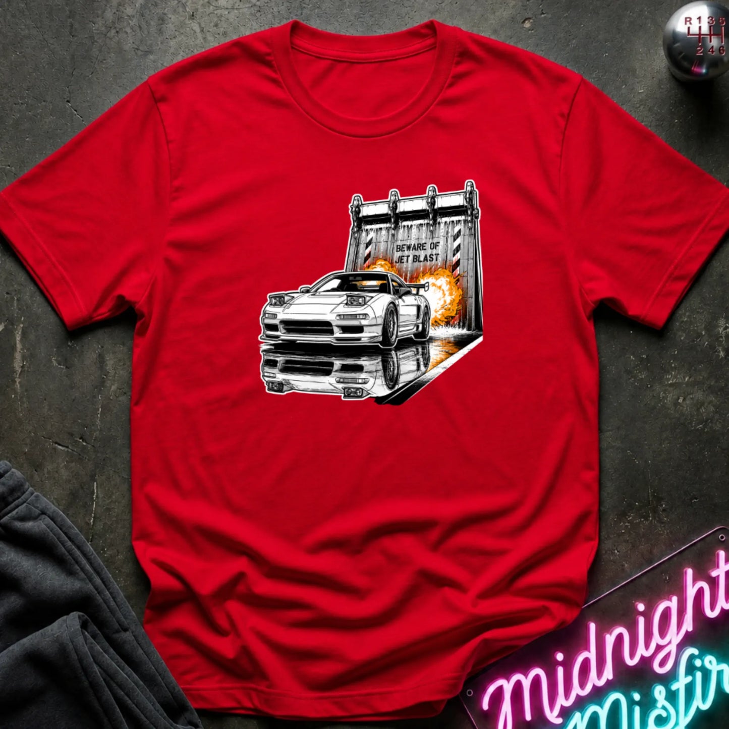 A red short-sleeve t-shirt laid flat on a dark textured surface, featuring a graphic print of a white sports car in front of a blast off area.