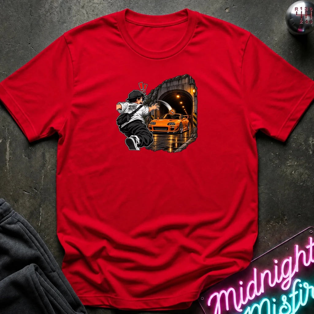 A red short-sleeve crewneck t-shirt laid flat on a dark textured surface. The center of the shirt features a graphic print of an anime character in a black and white outfit with a hat, next to a sports car 