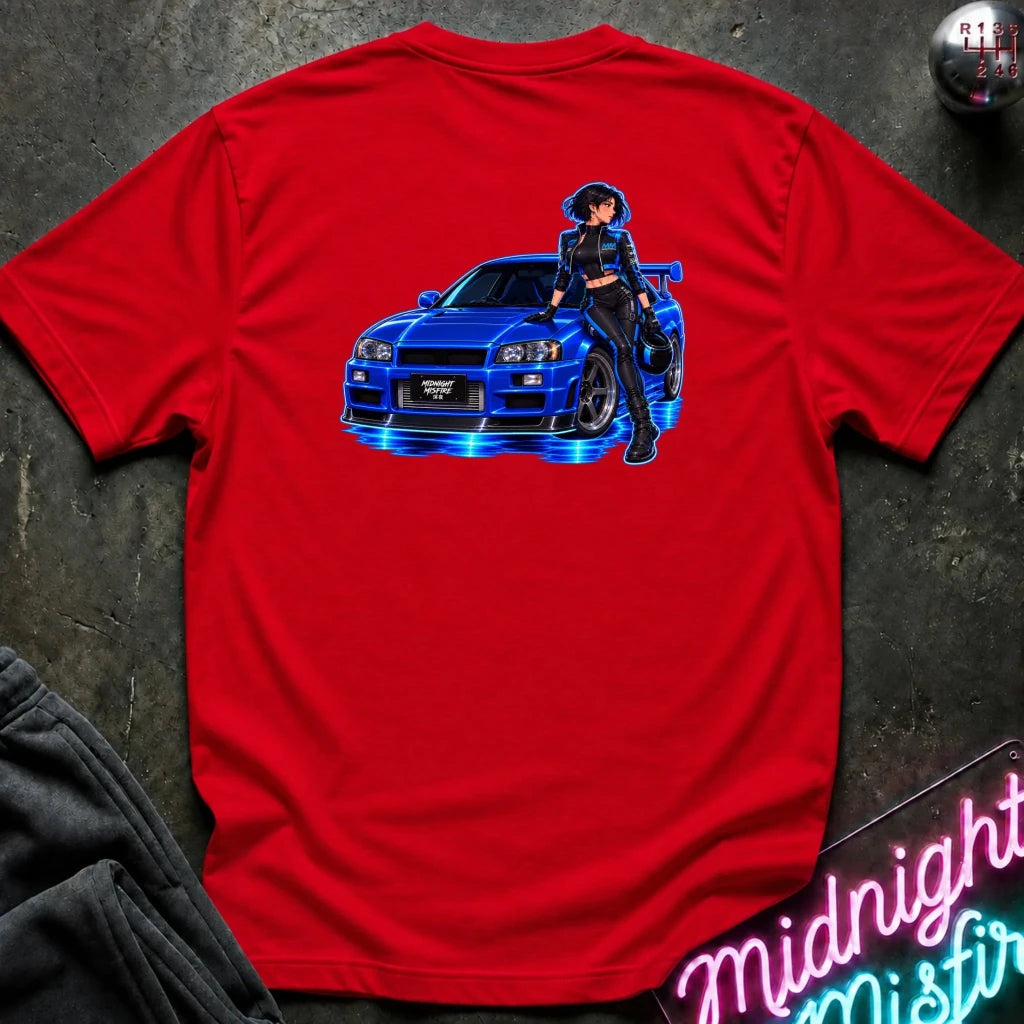A red short-sleeve t-shirt laid flat on a dark textured surface, featuring a graphic print of a blue sports car with glowing blue lights and an anime-style woman standing beside it. Partially visible props include dark pants in the lower left and a neon-style sign in the lower right.