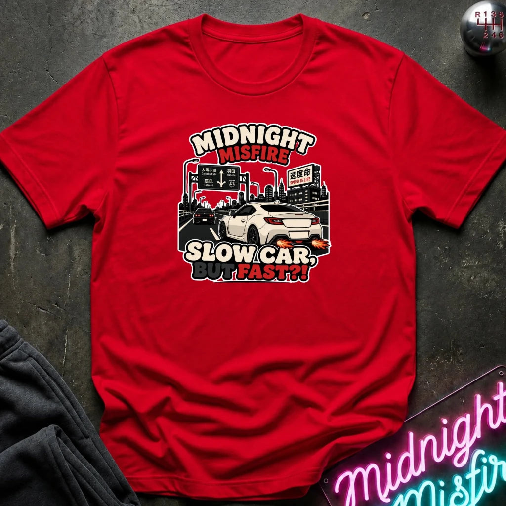 A red short-sleeve graphic t-shirt laid flat on a dark textured surface. The graphic features a white sports car and the text 'MIDNIGHT MISFIRE! SLOW CAR, FAST!!' in stylized lettering.