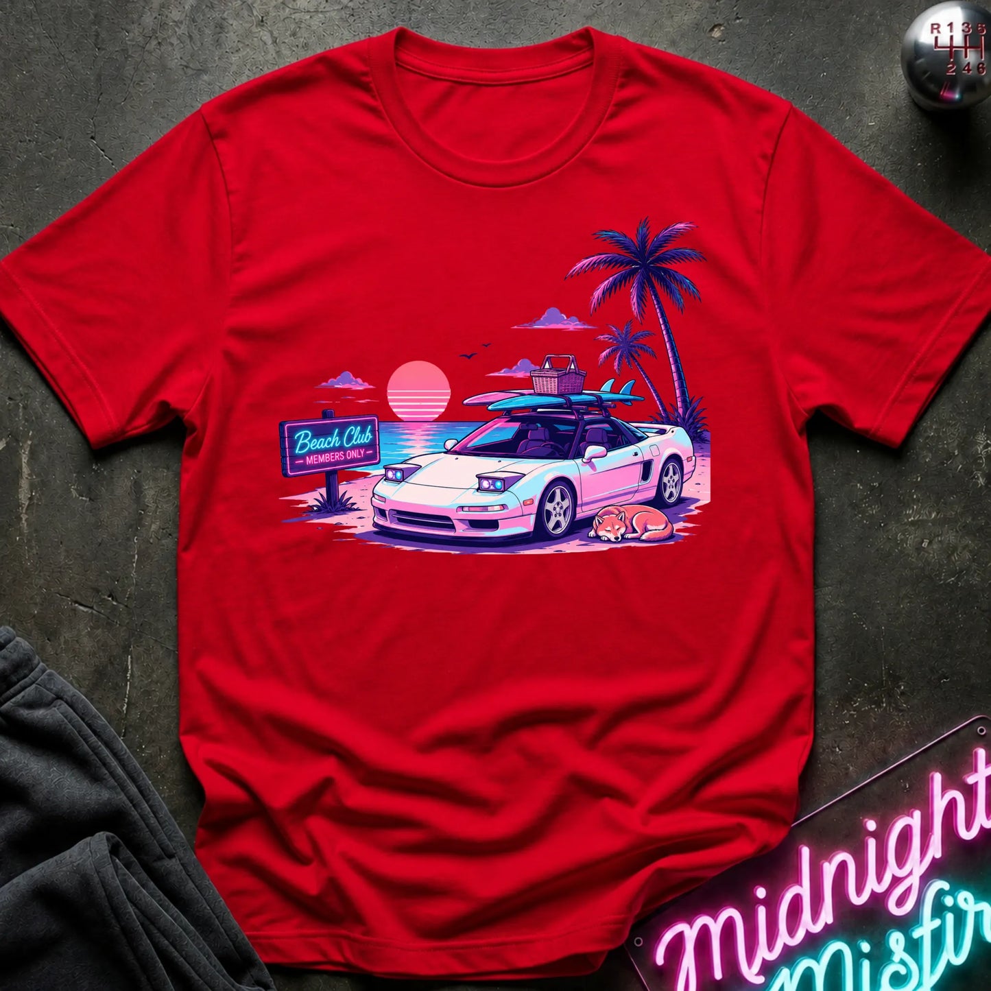 A red short-sleeve graphic t-shirt laid flat on a dark textured surface. The graphic features a retro sunset beach scene with a white sports car, palm trees, and a neon sign.