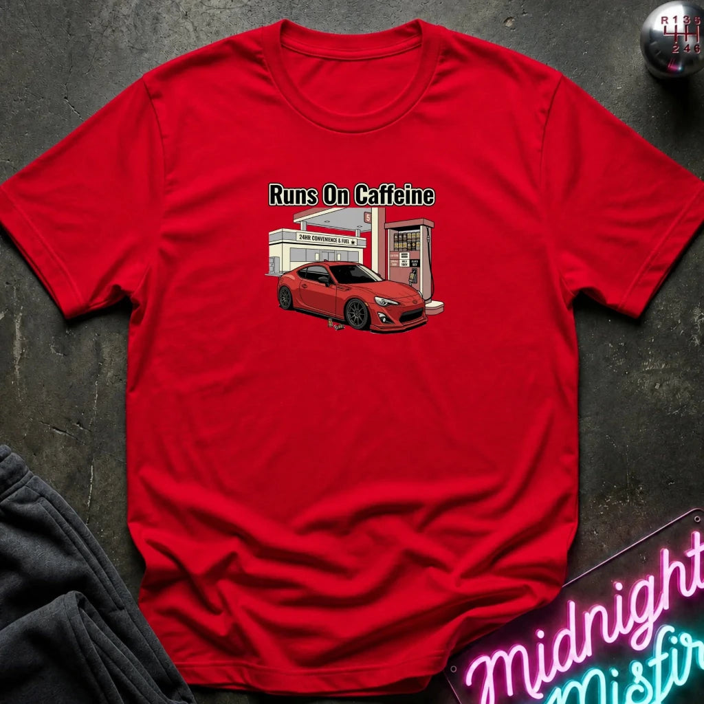 A red short-sleeve crewneck t-shirt laid flat on a dark textured surface. The shirt features a graphic print of a red sports car parked in front of a retro gas station with the text 'Runs On Caffeine' above it.