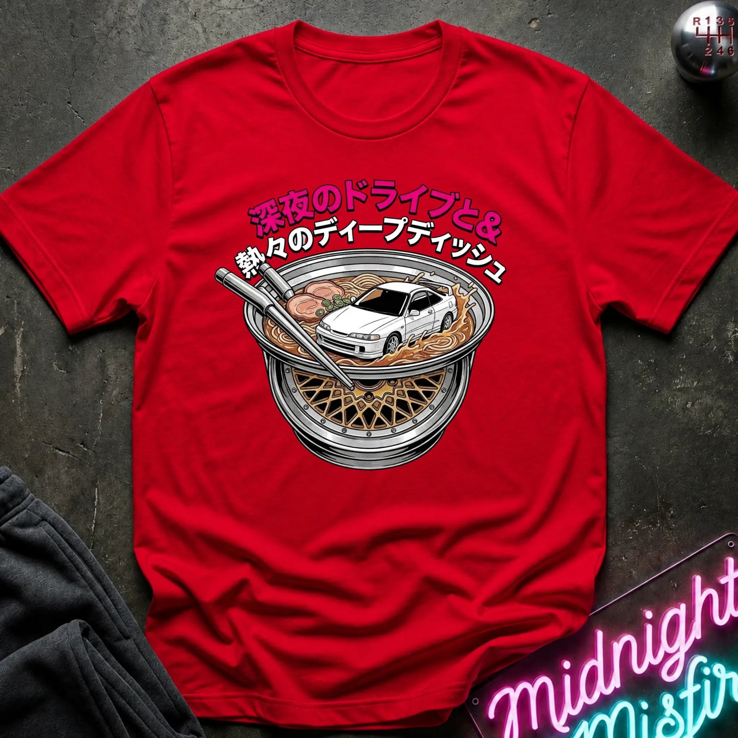A bright red short-sleeve graphic T-shirt laid flat on a dark textured surface. The shirt features a central illustration of a white sports car inside a ramen bowl with Japanese text above it.