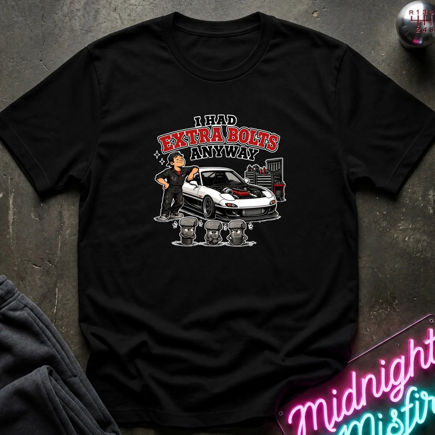 A black short-sleeve graphic t-shirt laid flat on a dark textured surface. The graphic features a cartoon of a man standing next to a white sports car with red detailing, with the text 'I HAD EXTRA BOLTS ANYWAY' in red and black.