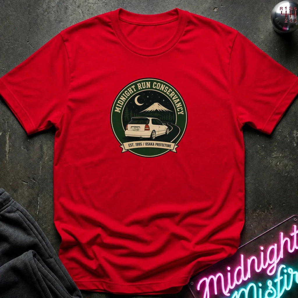 A red short-sleeve crewneck t-shirt laid flat on a dark textured surface. The center of the shirt features a circular graphic with a white SUV, a mountain, a crescent moon, and the text 'MIDNIGHT RUN CONSERVANCY' and 'EST.