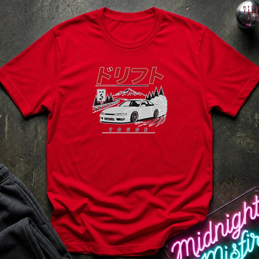 A red short-sleeve graphic T-shirt laid flat on a dark textured surface. The shirt features a white car illustration with Japanese text above and the word 'TOUGE' below.