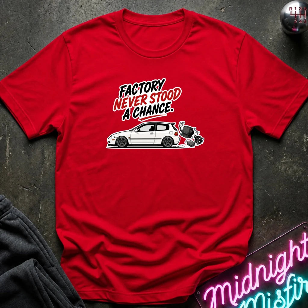 A red short-sleeve graphic T-shirt laid flat on a dark textured surface. The shirt features a white car illustration and the text 'FACTORY NEVER STOOD A CHANCE.