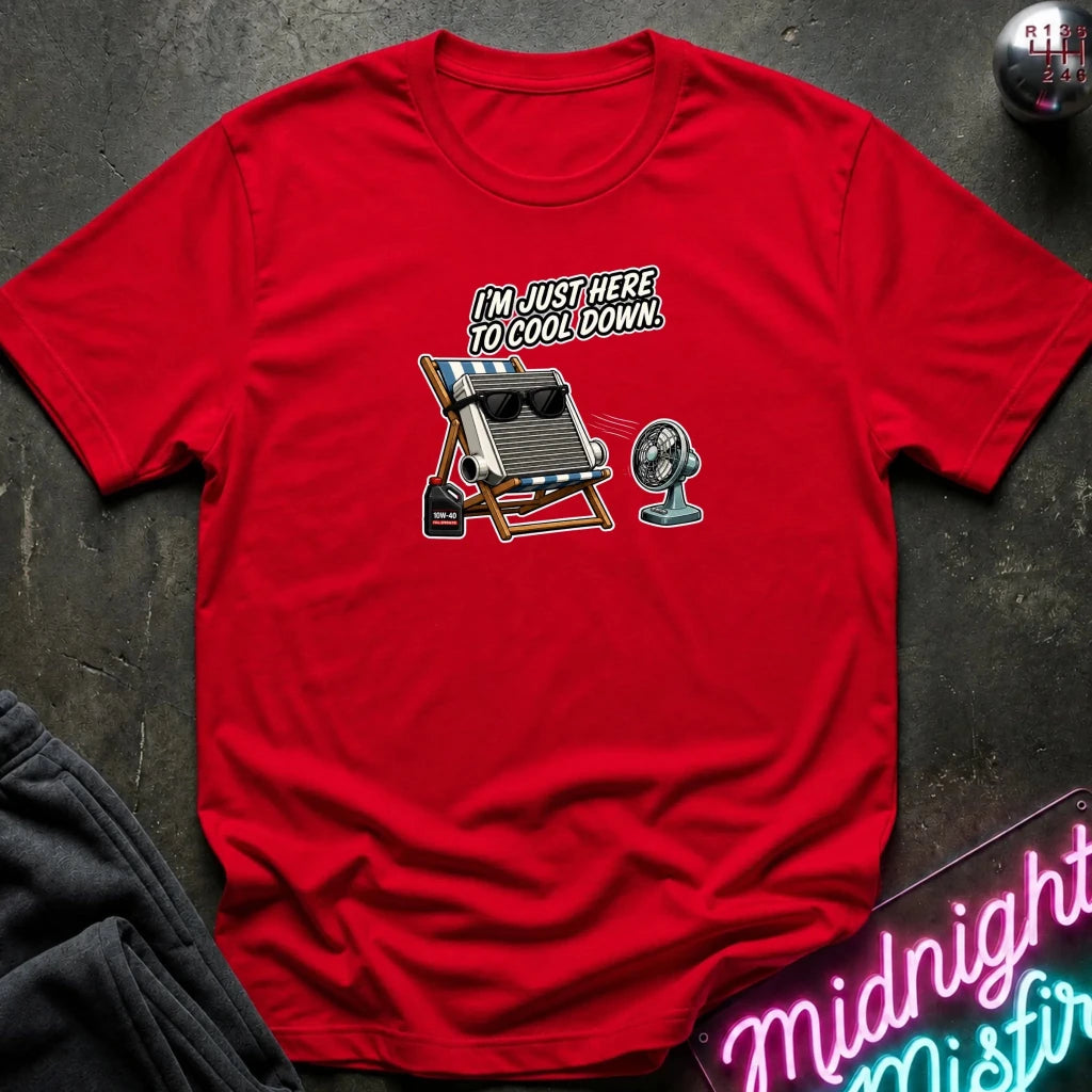 A red short-sleeve graphic t-shirt laid flat on a dark textured surface. The graphic features a vintage-style illustration of a beach chair with sunglasses, a small fan, and the text 'I'M JUST HERE TO COOL DOWN.
