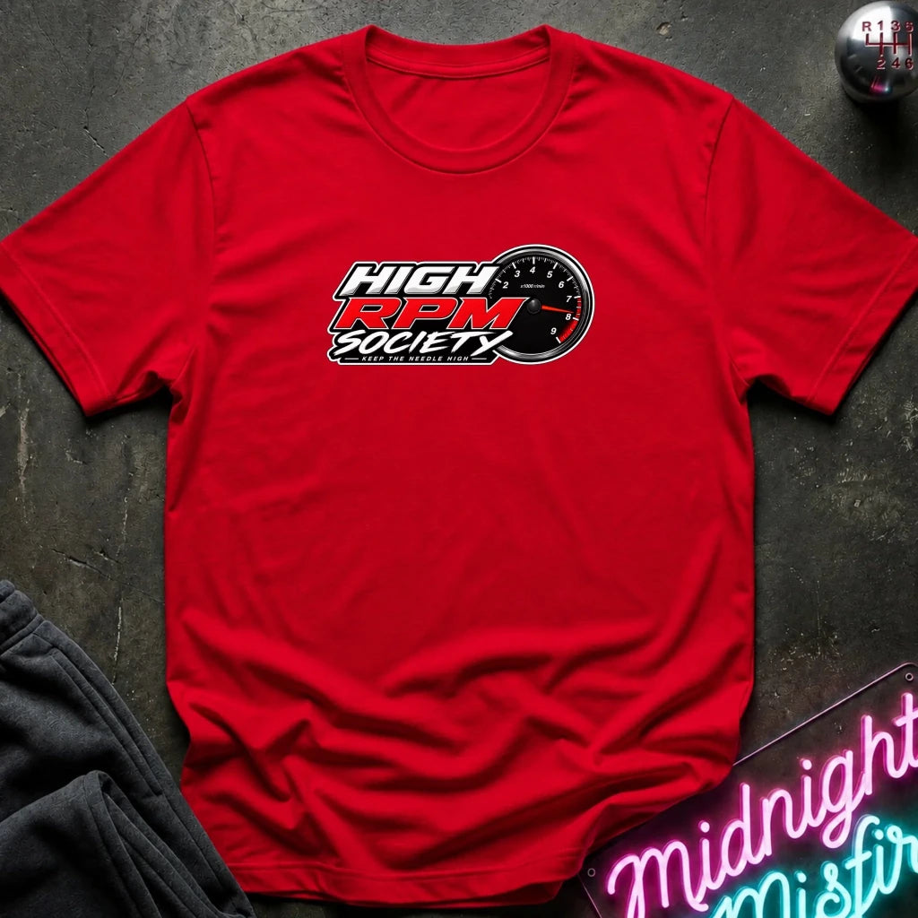 A red short-sleeve t-shirt laid flat on a dark textured surface, featuring a 'High RPM Society' logo printed on the chest. The shirt is styled with a dark gray sweatshirt in the corner and a neon sign in the foreground.