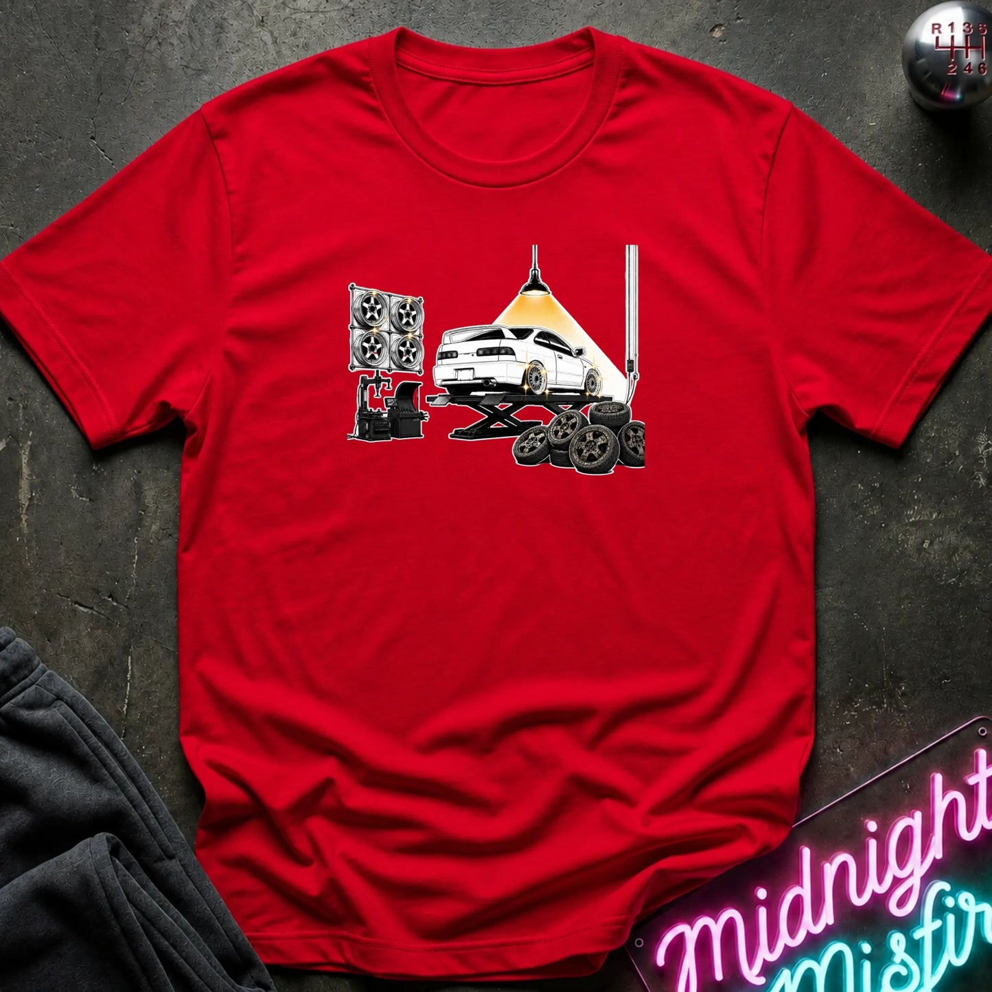 A red short-sleeve crewneck t-shirt laid flat on a dark textured surface. The shirt features a graphic print of a white car on a hydraulic lift with new wheels.