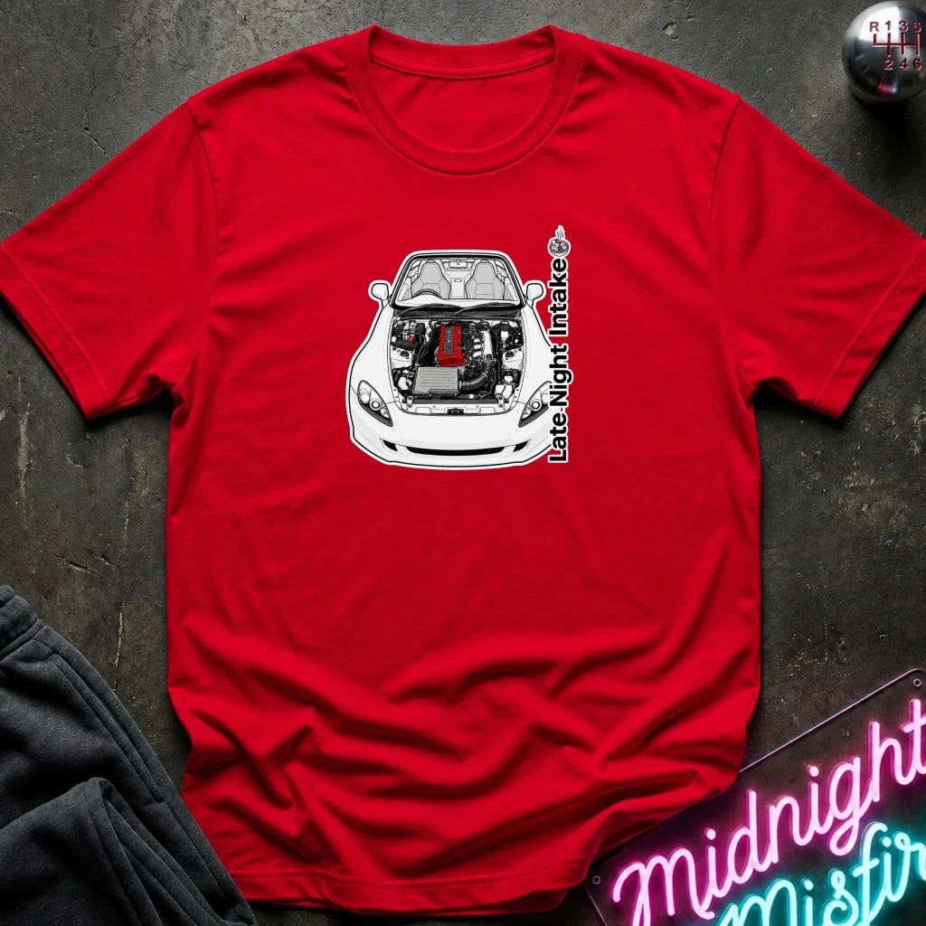 A red short-sleeve crewneck t-shirt laid flat on a dark textured surface. The shirt features a graphic print of a white sports car with its hood open, showing a red engine, with vertical text reading 'Late Night Intake'.