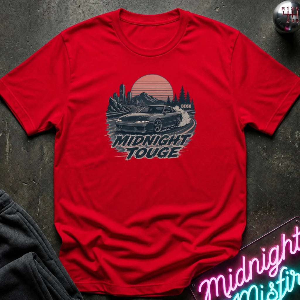 A red short-sleeve graphic T-shirt laid flat on a dark textured surface. The shirt features a retro-style circular print with a car drifting in front of mountains and trees, with the words 'MIDNIGHT TOUGHE' below.