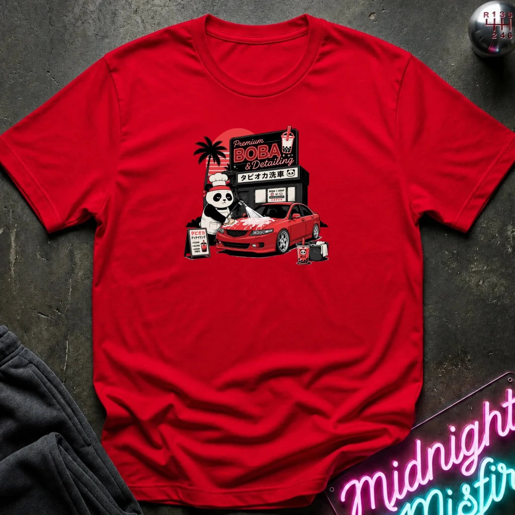 A red short-sleeve graphic t-shirt laid flat on a dark textured surface. The graphic features a cartoon panda wearing a hat next to a red sports car, with a sign reading 'Midnight Boba'.