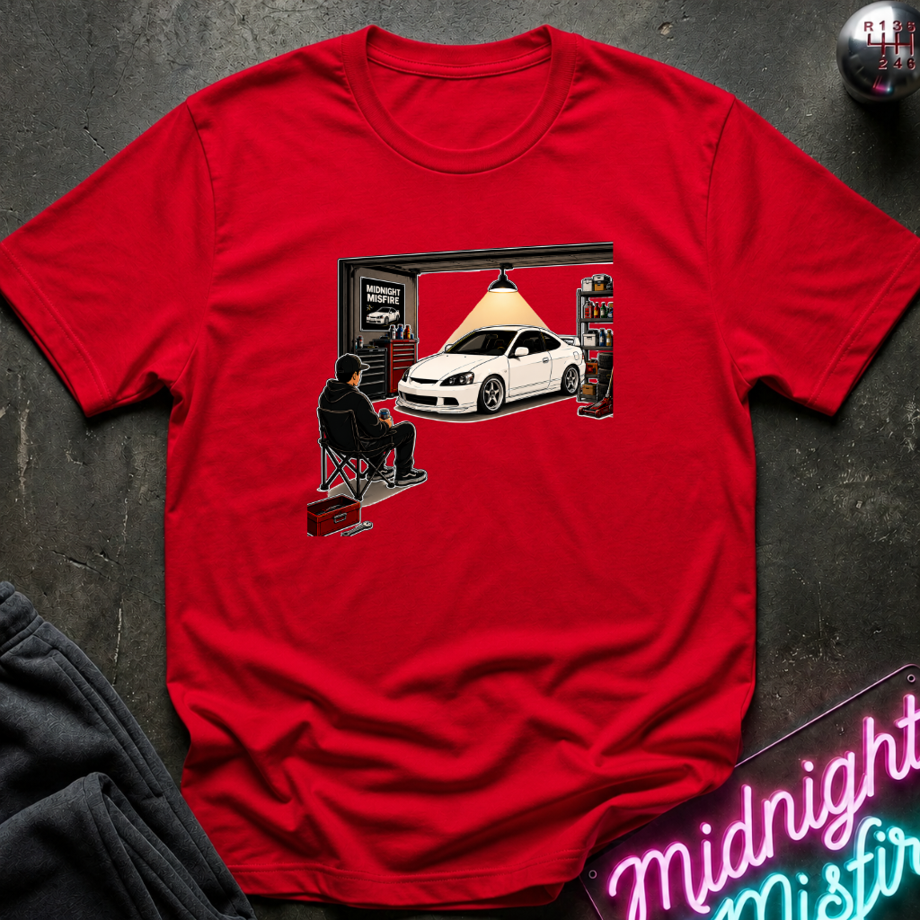 A red short-sleeve crewneck t-shirt with a graphic print of a white sports car under a spotlight in a garage, styled flat on a dark textured background with a neon sign and sweatpants partially visible.