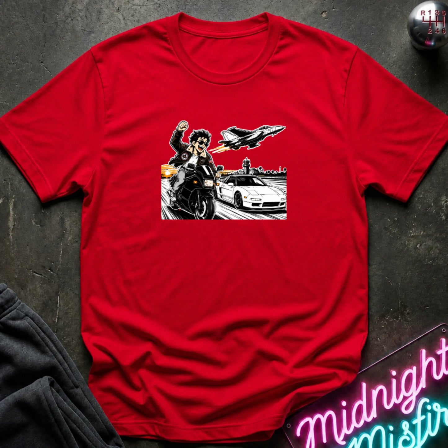A red short-sleeve crewneck t-shirt with a graphic print of a motorcycle racer, a sports car, and a jet plane, laid flat on a dark textured background with a neon sign and sweatpants partially visible.