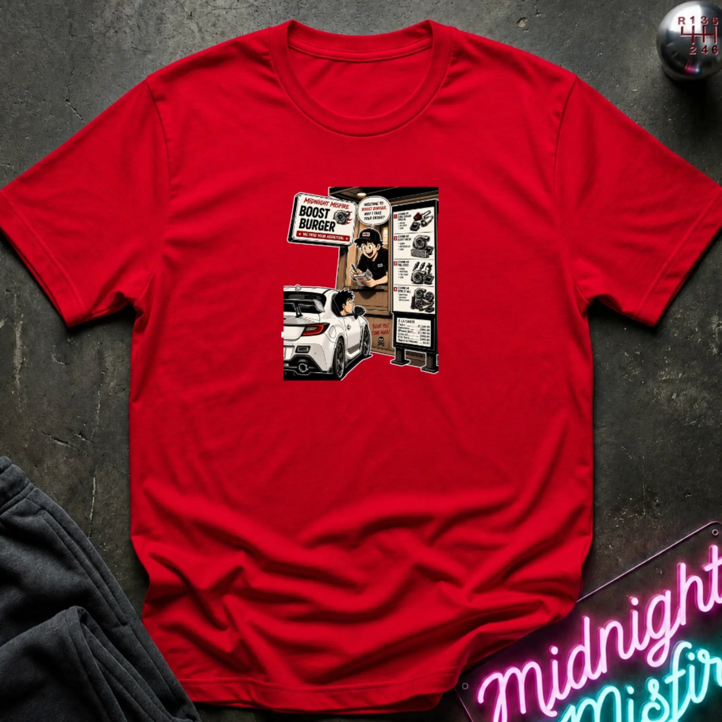 A red short-sleeve crewneck t-shirt laid flat on a dark textured surface, featuring a central graphic print of a street-food burger scene with a white car and signs. 