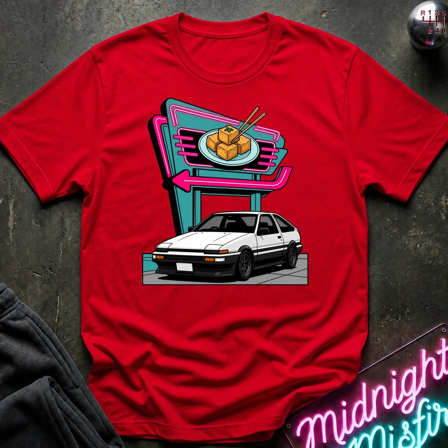 A red short-sleeve crewneck t-shirt featuring a retro-style graphic of a white sports car parked in front of a neon sign. The shirt is displayed flat on a dark textured surface with a dark sweatshirt and a neon sign partially visible as props.