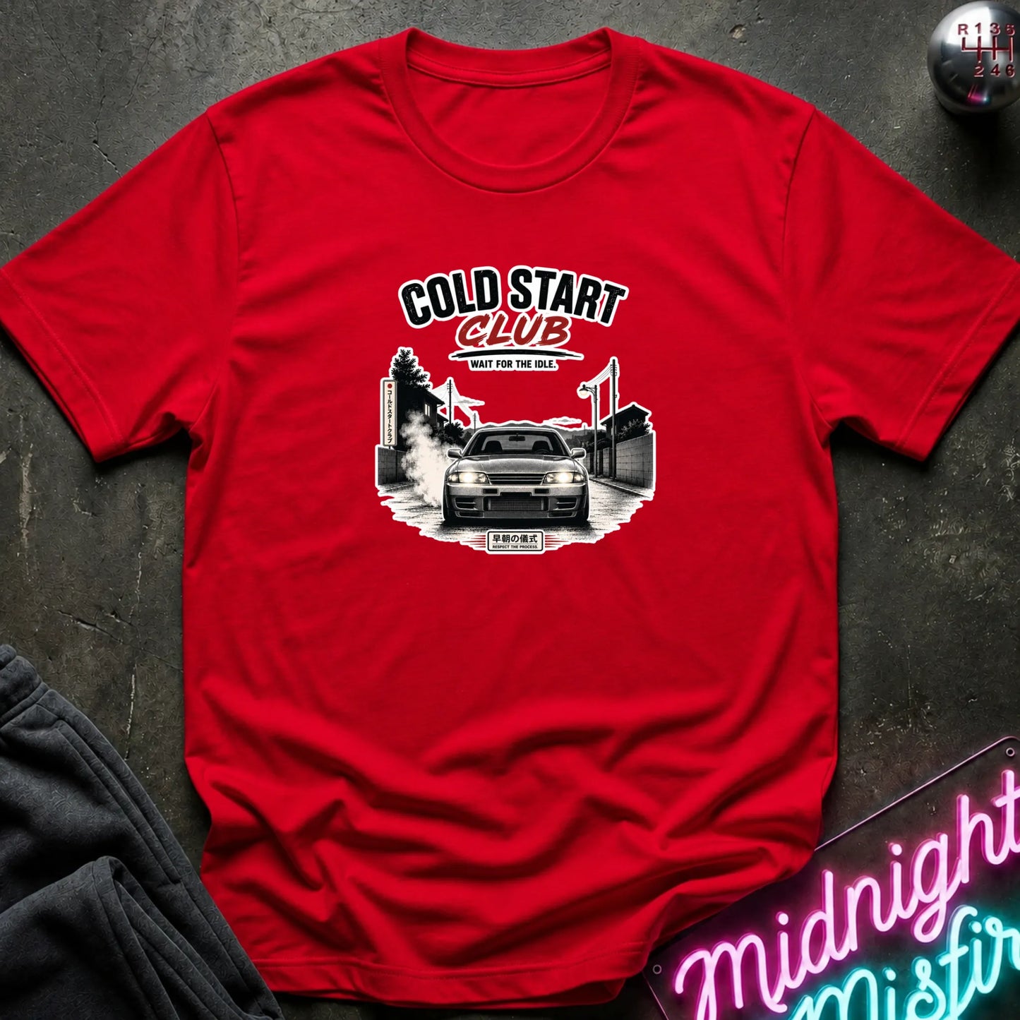 A red short-sleeve graphic T-shirt laid flat on a dark textured surface. The shirt features a black and white graphic of a car with the text 'COLD START CLUB'.