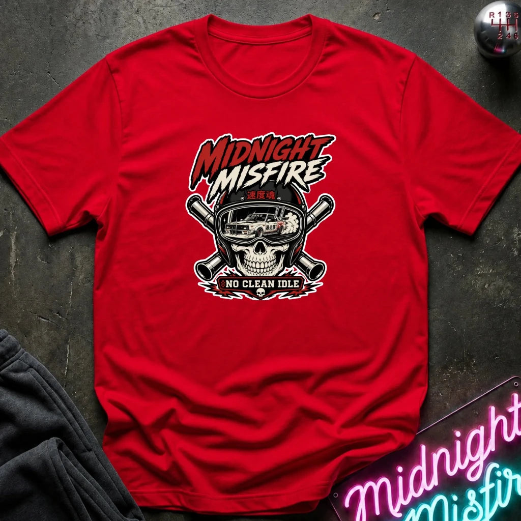 A red graphic t-shirt laid flat on a dark textured surface. The shirt features a central graphic of a skull wearing a racing helmet with crossed pipes behind it, and the text 'MIDNIGHT MISFIRE' and 'NO CLEAN IDLE'.