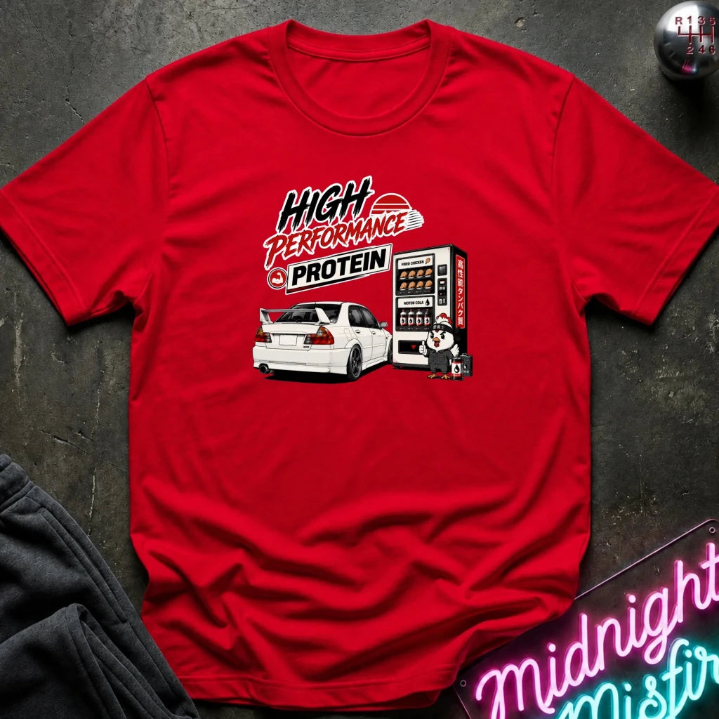 A red short-sleeve graphic T-shirt laid flat on a dark textured surface. The shirt features a printed design with the words 'HIGH PERFORMANCE PROTEIN' above a white sports car and a vending machine illustration.