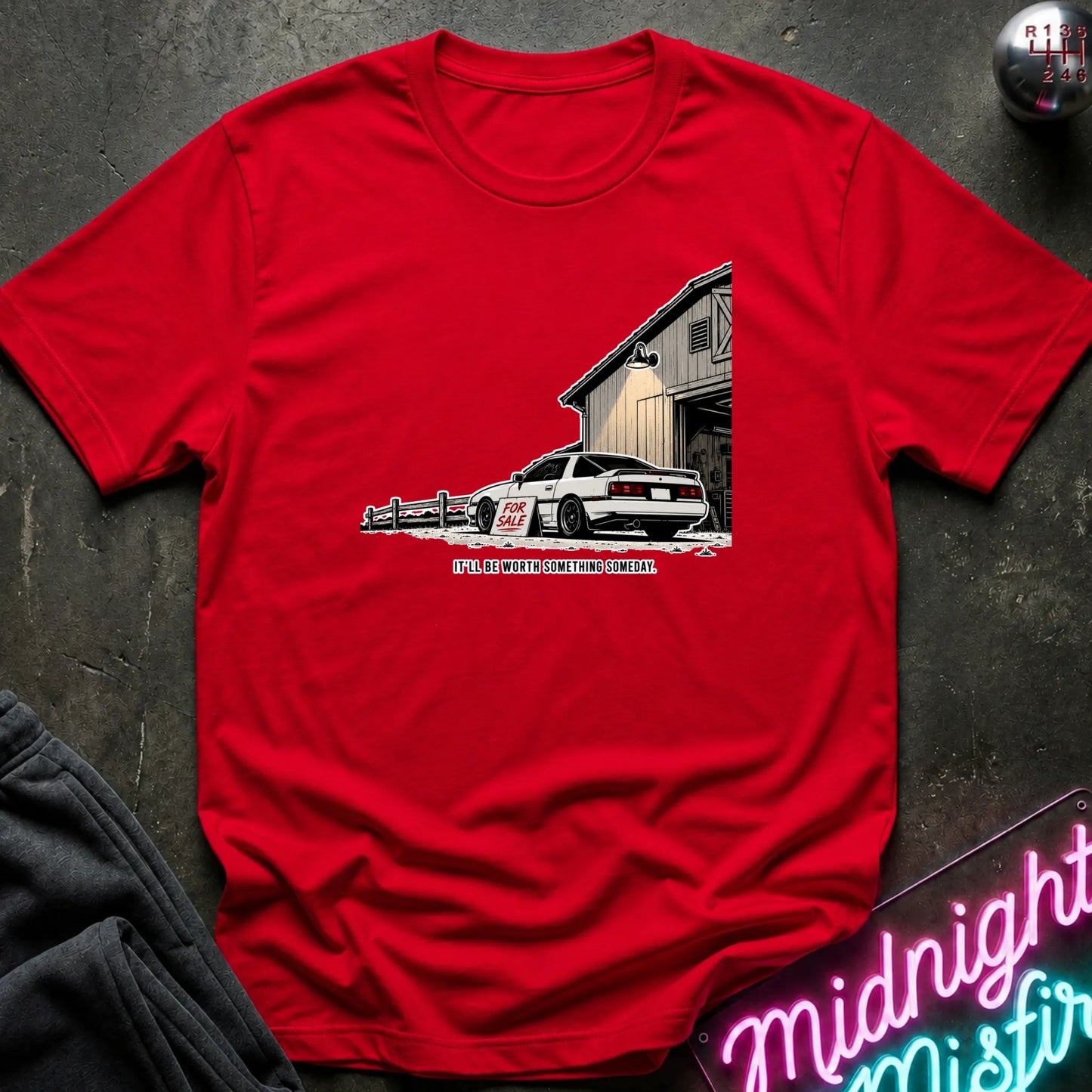 A red short-sleeve crewneck t-shirt laid flat on a dark textured surface. The shirt features a graphic print of a white race car parked in front of a barn.
