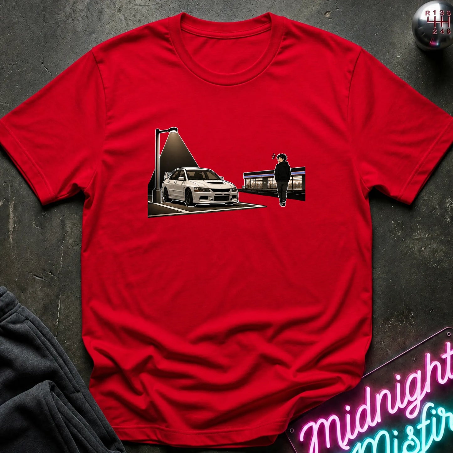 A red short-sleeve crewneck t-shirt laid flat on a dark textured surface. The shirt features a graphic print in the center depicting a white sports car under a spotlight next to a figure standing