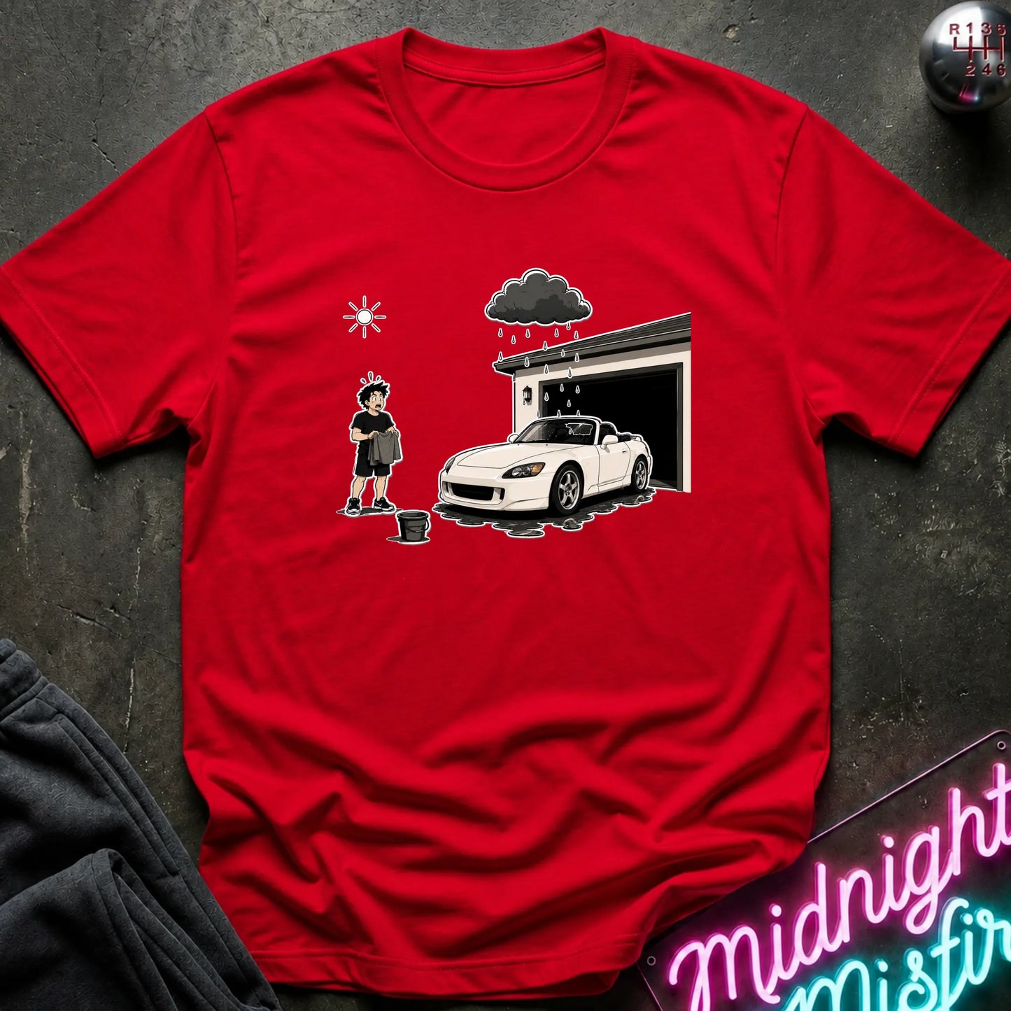 A red short-sleeve crewneck t-shirt featuring a cartoon graphic of a woman washing a white sports car in front of a garage under a dark stormy cloud. The shirt is displayed flat on a dark textured surface with a neon sign and dark sweatpants partially visible.