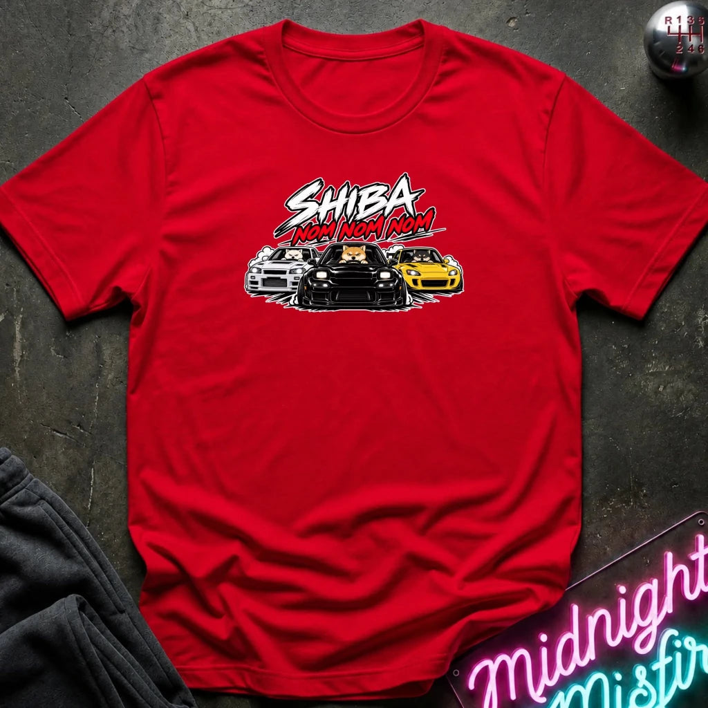 A red short-sleeve graphic t-shirt laid flat on a dark textured surface. The graphic features the text 'SHIBA NOM NOM NOM' in white and red, above an illustration of three cars (white, black, and yellow).